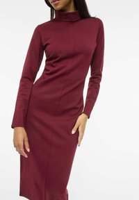 Ilga burgundiško vyno spalvos suknelė ilgomis rankovėmis, su aukštu kaklu, siaura linija, tekstūruotos medžiagos ir vertikalios siūlių detalės. Lygus apdaila, iki kelių ilgio.