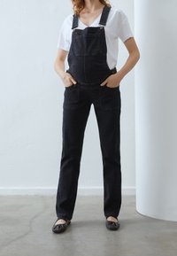 Svarta denimoverall med justerbara remmar, framfickor och avsmalnande ben, kombinerat med en vit T-shirt med V-hals och svarta ballerina skor.