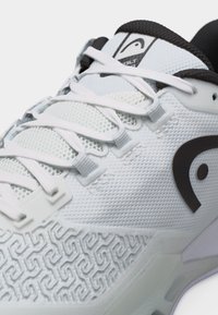 Λευκό αθλητικό παπούτσι με υφή στην πλευρική επιφάνεια, λευκά κορδόνια, μαύρη επένδυση εσωτερικά και μαύρο λογότυπο στο πλάι και στη γλώσσα.