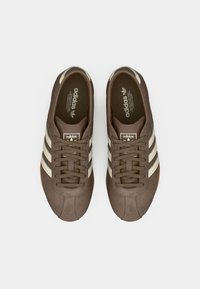 Bruna mockasneakers med tre krämvita ränder, texturerade skosnören och gummisulor, med en logotyp på tungan och innersulan.