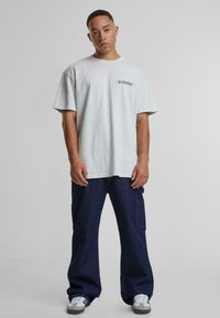 T-shirt gris à manches courtes avec un logo sur la poitrine, associé à un pantalon en denim bleu foncé ample et des baskets grises. Tenue simple et décontractée.