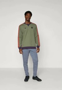 Green sweatshirt with red stripes on sleeves and a navy collar, featuring a round logo and paired with light blue joggers and black sneakers.