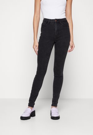 Vaqueros negros de cintura alta con corte ajustado, con cierre de botón, bolsillos frontales y una textura de mezclilla suave. Combinados con zapatos de color púrpura claro.