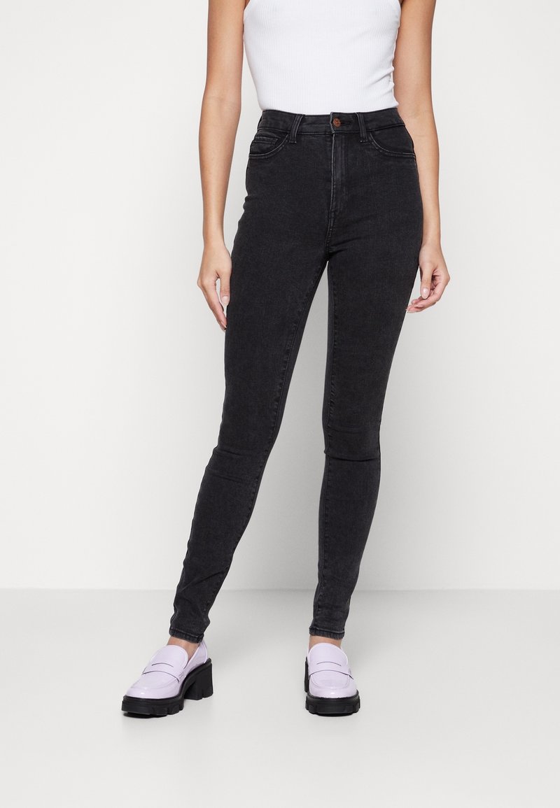 Vaqueros negros de cintura alta con corte ajustado, con cierre de botón, bolsillos frontales y una textura de mezclilla suave. Combinados con zapatos de color púrpura claro.