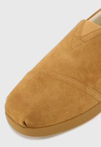 Zapato de ante marrón claro sin cordones, con punta redondeada, que presenta detalles de costura y una suela de goma suave. Su textura blanda mejora la comodidad.