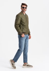 Olivgrön vadderad bomberjacka med sidizipper; kombinerad med ljusblå jeans och grå sneakers med vita detaljer och snören.