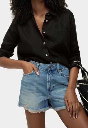 Mujer con camisa negra abotonada de manga remangada y pantalones cortos vaqueros azul claro desgastados, sosteniendo un bolso a rayas negro y blanco.