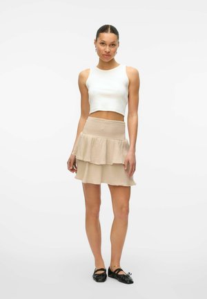 Frau mit einem Dutt, die ein weißes, kurzes Tanktop, einen beigen, schichtigen Minirock, schwarze Ballettschuhe und ein goldenes Fußkettchen trägt, vor einem weißen Hintergrund stehend.