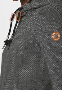 Donkergrijze hoodie met kleine witte stippen, zigzagstiksel bij de kraag, zwarte trekkoorden met metalen uiteinden en een ronde bruine patch op de mouw.