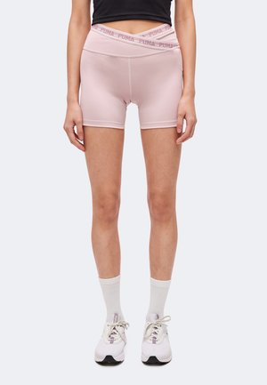 Femme portant un short de cycliste Puma rose pâle avec une taille croisée, des chaussettes blanches, des baskets blanches, et un top court noir, debout.