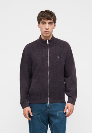 Giovane uomo che indossa un maglione viola scuro con zip integrale, collo e polsini a coste, abbinato a jeans blu, in piedi davanti a uno sfondo bianco uniforme.