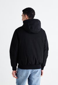 Chaqueta negra con capucha hecha de una tela suave, con un dobladillo y puños de canalé. Llevada sobre unos jeans de mezclilla azules, mostrando un ajuste relajado.