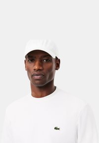 Cappellino da baseball bianco con texture liscia, dotato di visiera piatta. Indossato con una maglietta bianca unita, con un piccolo logo verde sul petto.