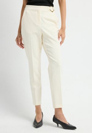 Slim-fit high-waist cream trousers with side pleats and a gold heart-shaped buckle, worn with black kitten heel shoes.