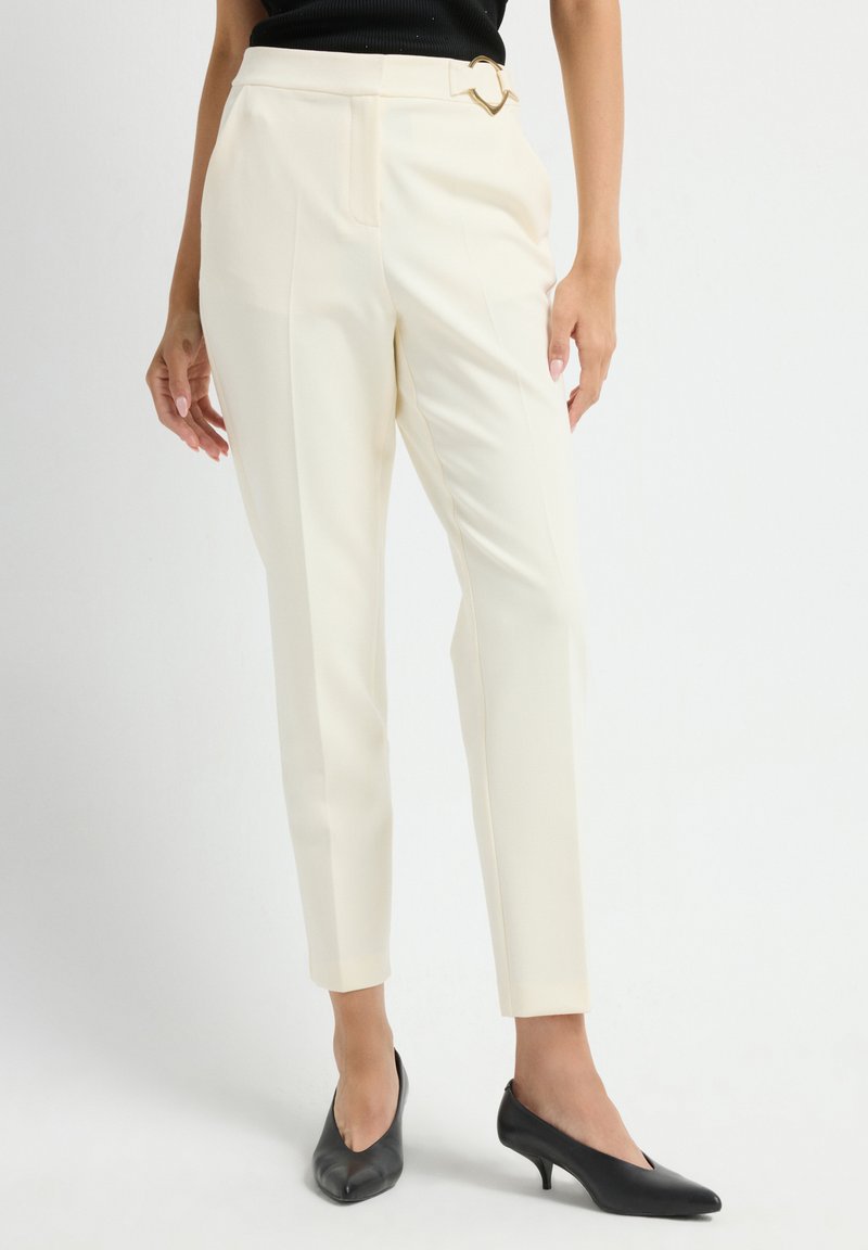 Slim fit krémové kalhoty s vysokým pasem, bočními záhyby a zlatou sponou ve tvaru srdce, nošené s černými botami na malém podpatku.