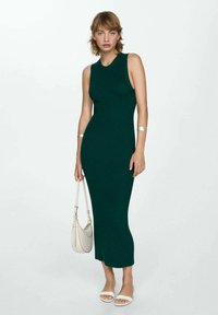 Abito a maglia senza maniche di un verde scuro, design aderente, lunghezza a metà polpaccio, texture liscia, abbinato a sandali bianchi e una borsa leggera.