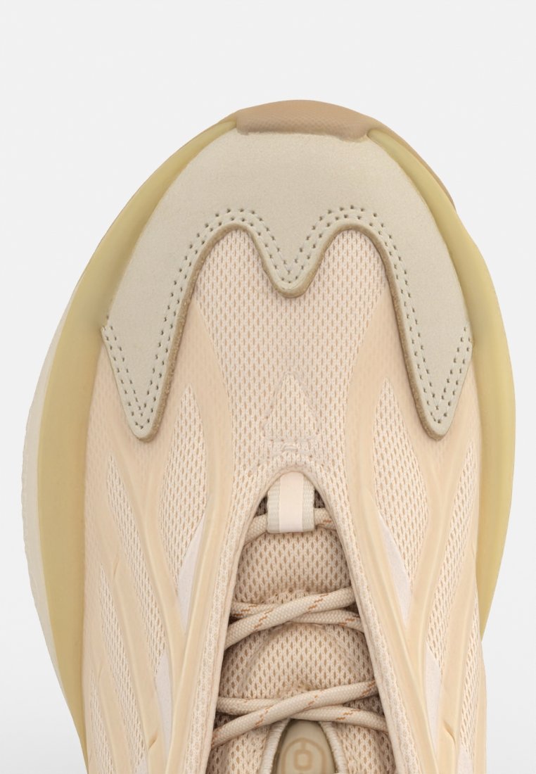 Zapatilla beige con parte superior de malla, puntera redondeada y diseño ondulado. Presenta cordones en el centro, costuras expuestas y una mediasuela texturizada.