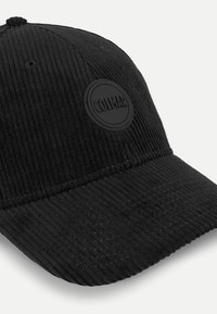 Črna korduro capica s krivim robom, teksturiranimi vertikalnimi črtami in krožno črno oznako na sprednji strani.