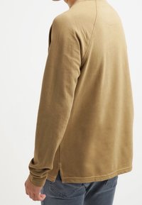 Haut à manches longues en tissu doux et texturé de couleur marron clair. Comprend des fentes latérales et un ourlet arrière arrondi pour plus de confort.