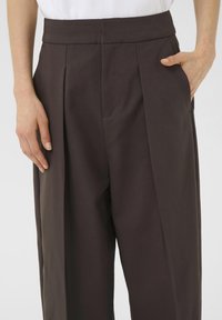 Pantaloni marroni a pieghe con una vestibilità su misura, caratterizzati da una texture liscia e tasche laterali, con un alto punto vita.