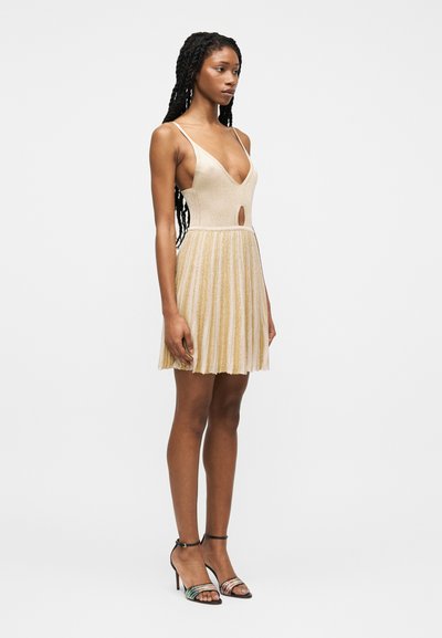 Vrouw met lang gevlochten haar, gekleed in een mouwloze beige gebreide jurk met geplooide rok en uitsnijdingsdetail, gecombineerd met open schoenen met hoge hakken.