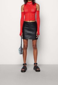 Punainen lyhythihainen neulepaita, jossa on pitkät hihat ja tekstuuri; yhdistetty mustiin mini-nahkahousuihin ja mustiin sandaaleihin. Kantaa raitakuvioista laukku.