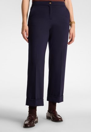 Donna che indossa pantaloni blu navy su misura con orli risvoltati, calzini marroni, mocassini in pelle marrone scuro e un braccialetto d'oro al polso destro.