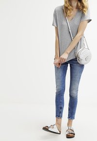 Gray short-sleeve shirt, fitted blue jeans with side zippers, and metallic sandals. White crossbody bag with quilted pattern.