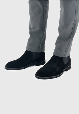 Zwarte suede Chelsea-laarzen met elastische zijkanten, ronde neus en lage hak, gecombineerd met grijze geklede broek.