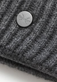 Gray ribbed knit beanie with a circular metallic tag embossed with "PEARLWOOl" and needle motifs, featuring a soft, textured finish.