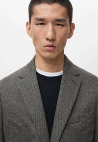 Grau kariertes Blazer mit Revers und Tasche, über einem schwarzen Pullover mit weißem Kragen getragen. Glatte Textur, taillierte Passform.