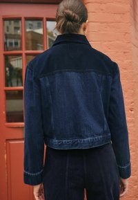 Mørk denimjakke med cropped skjæring, der har en kordfløjlskrave, synlig syning og sat mod en murstensbaggrund.