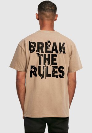 Beige t-shirt med korte ærmer, der har fed sort tekst på ryggen, som siger "BRYDE REGULERNE" med en slidt, revnet effekt.