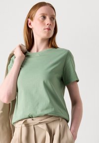 T-shirt in cotone verde chiaro con collo girocollo, maniche corte e un dettagliato logo discreto sul petto, abbinata a pantaloni beige a vita alta.