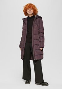Lila Daunenjacke mit Reißverschluss, Kapuze und horizontalem Steppmuster. Getragen über einem schwarzen Rollkragenpullover und kombiniert mit schwarzen Hose.