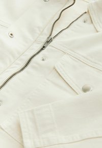 Gros plan sur une veste en denim blanc cassé avec une fermeture éclair argentée, des boutons-pression et une poche poitrine à rabat.
