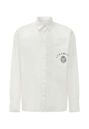 Camicia bianca a maniche lunghe con bottoni, colletto a punta e taschino frontale con stemma nero e logo testuale.