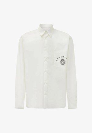 Camicia bianca a maniche lunghe con bottoni, colletto a punta e taschino frontale con stemma nero e logo testuale.