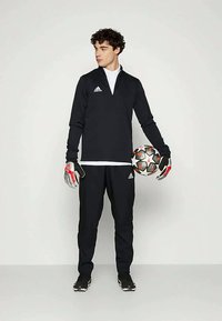 Fekete sportos melegítő szett hosszú ujjal, fehér trikó, kesztyűk és egy focilabdával. Az ingeden és a nadrágon Adidas logó található.