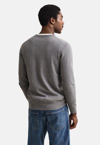 Mężczyzna zwrócony tyłem, ubrany w jasnoszary sweter z długim rękawem i niebieskie dżinsy na jednolitym białym tle.