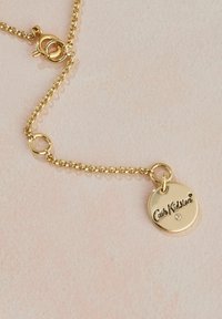 Kuldne kaelakee, millel on ümmargune ripats, millel on graveeritud tekst "Cath Kidston" ja väike sisse tunginud kivi. Lihtne disain ja sile tekstuur.