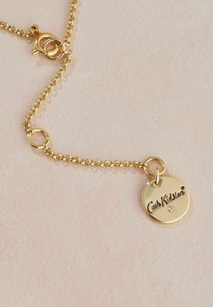 Collana in oro con un pendente circolare che presenta il testo inciso "Cath Kidston" e una piccola pietra incastonata. Design semplice e texture liscia.