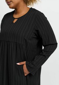 Schwarzes Langarmkleid mit vertikaler Ripptextur, einem Schlüsselloch-Ausschnitt und Seitentaschen. Lässige Passform mit fließender Silhouette.