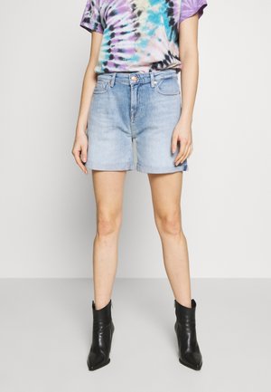 Personne portant un short en jean bleu clair, un t-shirt tie-dye à manches courtes et des bottines noires, debout devant un fond uni.