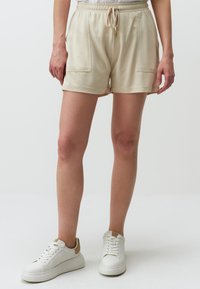 Beige katoenen shorts met een elastische tailleband, trekkoorddetails en twee voorzakken, gedragen met witte sneakers met tan accenten.