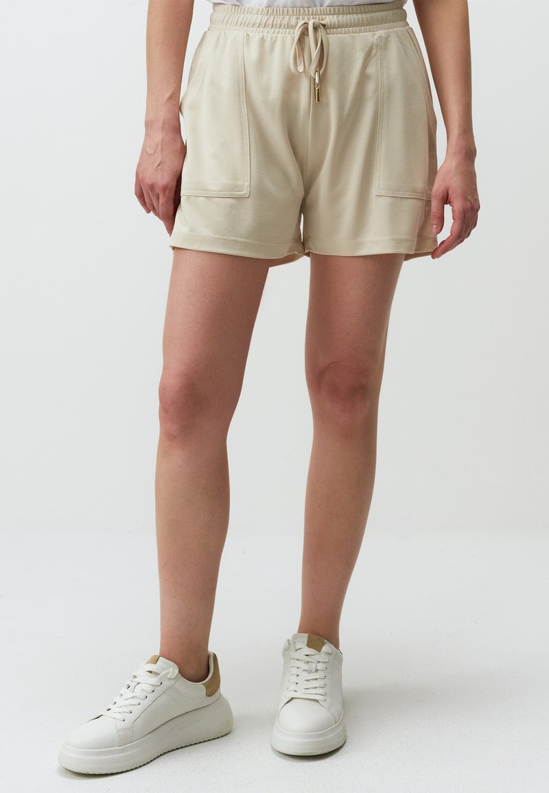 Beige katoenen shorts met een elastische tailleband, trekkoorddetails en twee voorzakken, gedragen met witte sneakers met tan accenten.
