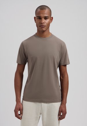 Man with close-cropped hair wearing a taupe short-sleeve t-shirt and off-white pants, standing against a plain light gray background.