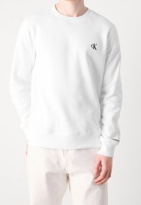 Vit sweatshirt med rund hals, långa ärmar och ribbade muddar. Har en liten svart logotyp på bröstet och text på ärmsluten.