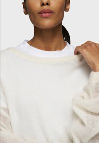 Maglione bianco leggero a maglia con maniche leggermente trasparenti e scollo ampio, indossato sopra una maglietta bianca a girocollo. Texture liscia.