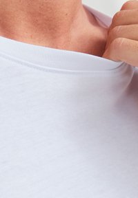 T-shirt en coton blanc avec un col rond, montrant une texture lisse et des détails de couture près du col, partiellement tiré loin du cou.